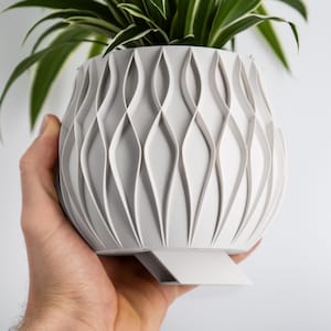 Puede incluir: Maceta de cerámica blanca con un diseño único, ondulado y texturizado. La maceta tiene una base plana con un pequeño borde.
