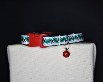 May include: A white cat collar with a red buckle and a small red bell. The collar features a pattern of green holly leaves and red berries. The collar is displayed on a neutral-colored surface against a dark background.