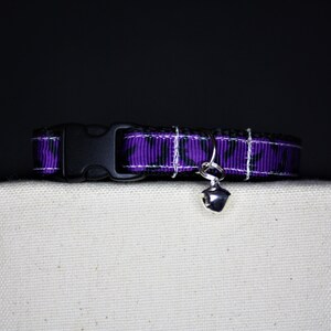 May include: A cat collar featuring a purple and black pattern, a black plastic buckle, and a silver bell. The collar is presented on a white surface, set against a dark backdrop.