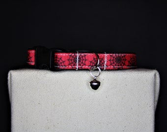 May include: Red and black patterned cat collar with a black plastic buckle and a silver bell charm. The collar is displayed on a neutral-colored surface against a black background. The collar's pattern resembles a spider web.