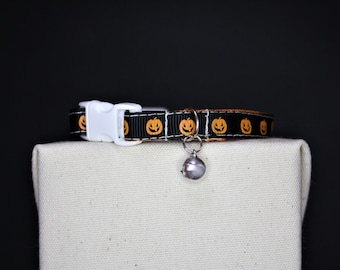 May include: Black cat collar with orange jack-o'-lanterns. The collar has a white plastic buckle and a silver bell attached. The collar is displayed on a beige surface against a black background. A festive accessory for pets.