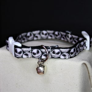 May include: A black and white patterned cat collar featuring a repeating design of a character's face. The collar has a white plastic buckle and a silver-colored bell. The collar is resting on a beige surface.