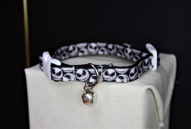 May include: A black and white patterned cat collar featuring a repeating design of a character's face. The collar has a white plastic buckle and a silver-colored bell. The collar is resting on a beige surface.