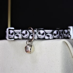 May include: A black and white cat collar featuring a repeating pattern of Jack Skellington's face. The collar has a white plastic buckle and a silver bell attached. The collar is displayed on a white surface against a dark background.