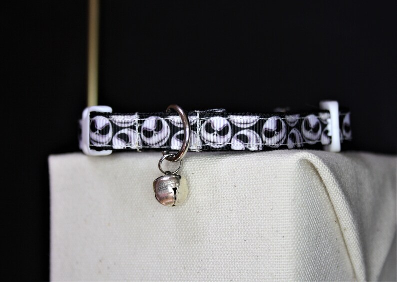 May include: A black and white cat collar featuring a repeating pattern of Jack Skellington's face. The collar has a white plastic buckle and a silver bell attached. The collar is displayed on a white surface against a dark background.