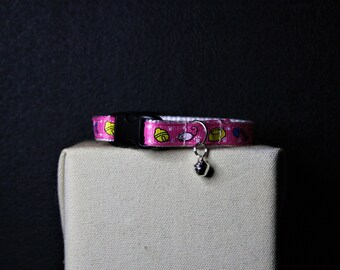 May include: A pink cat collar with a black buckle and a silver bell. The collar is decorated with a repeating pattern of yellow cat food bowls, yellow bells, and blue fish on a pink background.