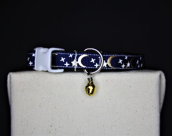 Op de afbeelding: Een marineblauwe kattenhalsband met een wit en gouden maan- en sterrenpatroon en een gouden belhanger.