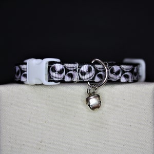 May include: A black and white cat collar featuring a repeating Jack Skellington face pattern. The collar has a white plastic buckle and a silver bell charm. The collar is displayed on a neutral-colored surface against a black background.