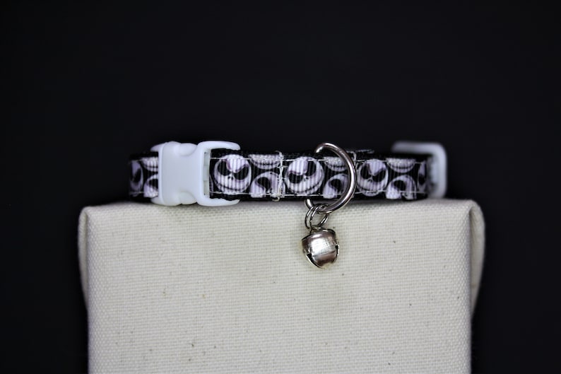 May include: A black and white cat collar featuring a repeating Jack Skellington face pattern. The collar has a white plastic buckle and a silver bell charm. The collar is displayed on a neutral-colored surface against a black background.