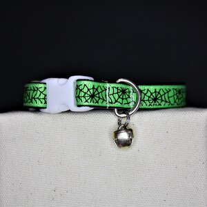 May include: A green cat collar with a black spiderweb design. The collar has a white plastic buckle, a silver metal D-ring, and a small silver bell. The collar is displayed on a white surface against a black background.