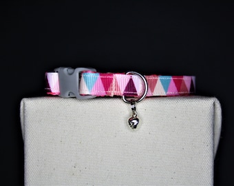 May include: A pink, blue, and white patterned cat collar with a gray plastic buckle and a silver bell charm. The collar features a repeating triangle design and is displayed on a neutral-colored surface against a black background.