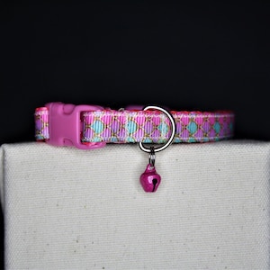 May include: A pink cat collar featuring a repeating pattern of gold, teal, and purple diamonds. The collar has a pink plastic buckle and a small pink bell attached with a silver ring. The collar is displayed on a white surface against a black background.