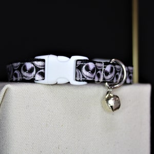 May include: A black and white patterned cat collar featuring a repeating design of a character's face. The collar has a white plastic buckle and a small silver bell attached. The collar is resting on a beige surface.