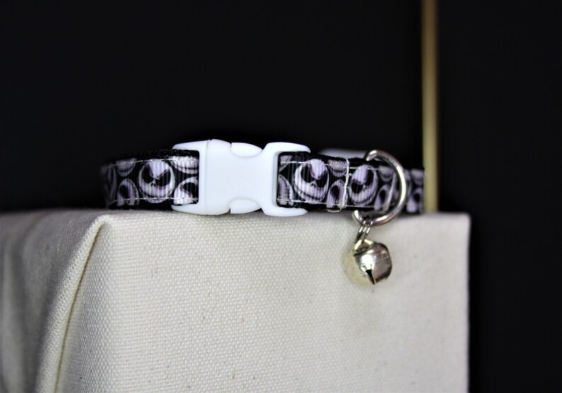 May include: A black and white patterned cat collar featuring a repeating design of a character's face. The collar has a white plastic buckle and a small silver bell attached. The collar is resting on a beige surface.