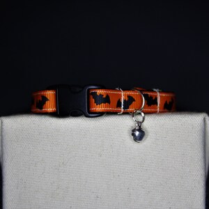 May include: An orange and black cat collar featuring a bat design, with a black plastic buckle and a small silver bell. The collar is displayed on a neutral surface against a dark backdrop, perfect for Halloween.