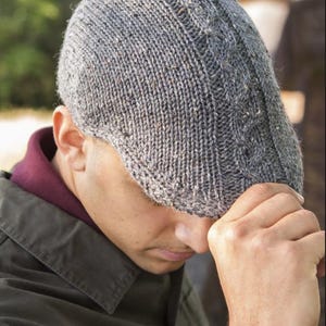 May include: A gray knitted flat cap with a textured pattern. The cap is made of a speckled yarn and has a classic, rounded shape. The image includes the text "PDF Knitting Pattern Download!"