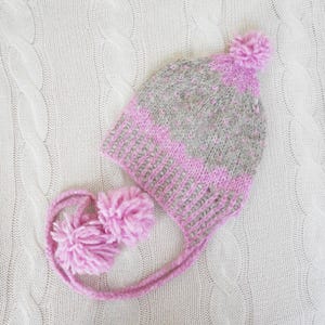 Puede incluir: Un gorro de punto en tonos rosa y gris, con un pompón en la parte superior y orejeras con dos pompones adicionales. El gorro está hecho de un hilo suave y texturizado, ideal para el invierno. Se muestra sobre una superficie de punto de cable crema.