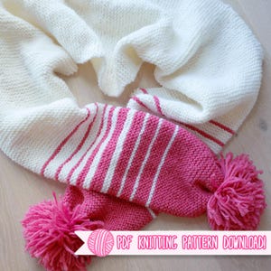 Könnte beinhalten: Ein cremefarbener Strickschal mit rosa und weißen Streifen und rosa Bommeln. Der Schal ist aus einem weichen, strukturierten Garn gefertigt. Der Text "PDF Knitting Pattern Download!" ist sichtbar.