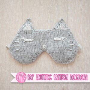 May include: A gray knitted sleep mask in the shape of a cat's face, featuring white accents for the ears, eyes, and whiskers. The mask has a soft, textured appearance and is designed to cover the eyes. Text on a pink banner reads "PDF KNITTING PATTERN DOWNLOAD!"