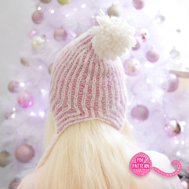May include: A pink and white striped knit hat with a large white pom-pom on top. The hat has ear flaps and is set against a blurred background of a white Christmas tree with pink ornaments. The hat is a PDF pattern.