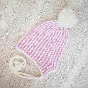 May include: A knitted winter hat with a pink and white striped pattern. The hat has ear flaps, a white tassel, and a large white pom-pom on top. The hat is lying on a cream-colored knitted blanket.