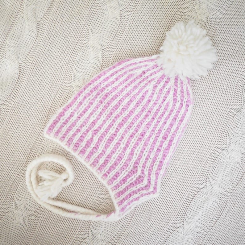 May include: A knitted winter hat with a pink and white striped pattern. The hat has ear flaps, a white tassel, and a large white pom-pom on top. The hat is lying on a cream-colored knitted blanket.