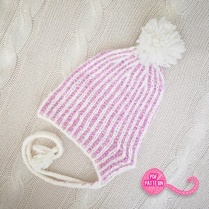 May include: A knitted winter hat with a striped pattern in white and light pink. The hat features a large white pom-pom on top and ear flaps with tassels. The hat is lying on a cream-colored knitted surface.