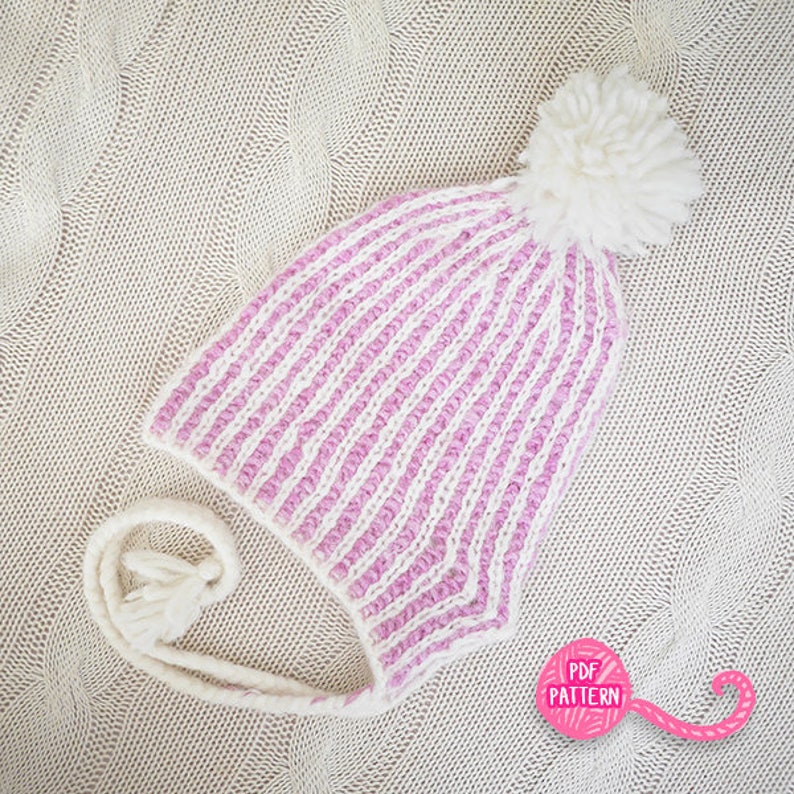 May include: A knitted winter hat with a striped pattern in white and light pink. The hat features a large white pom-pom on top and ear flaps with tassels. The hat is lying on a cream-colored knitted surface.