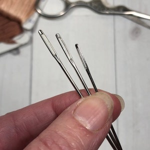 May include: Three silver sewing needles of different sizes, held in a hand.