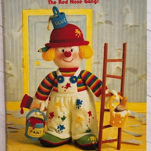 Könnte beinhalten: Buchcover "Jean Greenhowe's Knitted Clowns" mit einem gestrickten Clown mit rotem Hut, gestreiftem Oberteil und Latzhose. Der Clown hält einen Farbeimer und einen Pinsel. Der Text "The Red Nose Gang!" ist ebenfalls sichtbar.