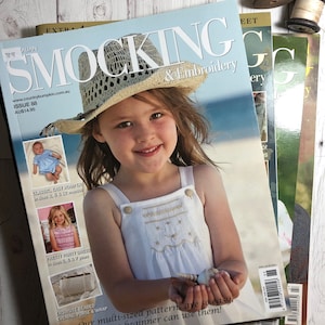 Puede incluir: Una pila de revistas, la superior con una niña vestida con un vestido blanco y un sombrero de paja. La revista se titula "SMOCKING & Embroidery". Otras revistas son visibles en el fondo, con texto e imágenes.