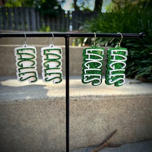 May include: Earrings with the word "EAGLES" in green glitter on a white background. The earrings are hanging from a black metal stand. The earrings have a rectangular shape.