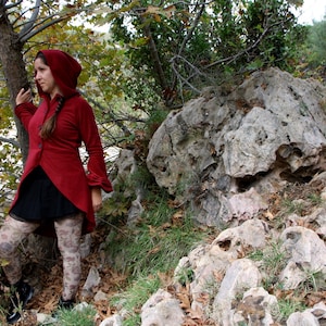 Può includere: Una donna indossa una giacca rossa con cappuccio, una gonna nera e leggings a fantasia in piedi in una zona rocciosa e boscosa.