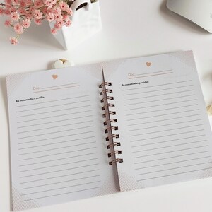 May include: An open spiral bound journal with a white background and a delicate pink and white pattern on the cover. The journal has lined pages and the words "Dia:" and "Mis pensamientos y sue&ntilde;os" are printed on the pages.