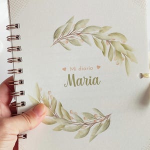 May include: A white journal with a spiral binding and a gold ribbon. The cover features a watercolor illustration of green leaves and pink flowers. The text "Mi diario Maria" is printed on the cover.