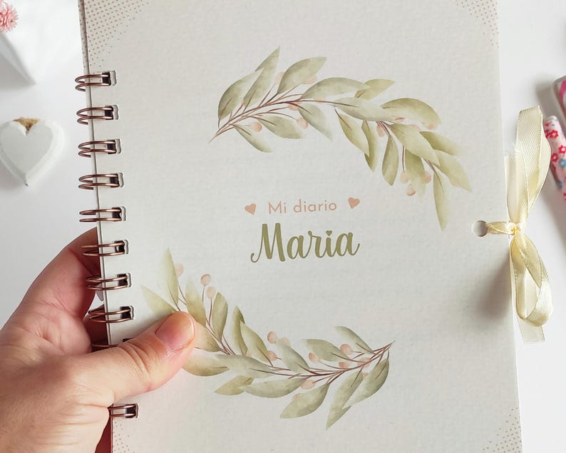 May include: A white journal with a spiral binding and a gold ribbon. The cover features a watercolor illustration of green leaves and pink flowers. The text "Mi diario Maria" is printed on the cover.