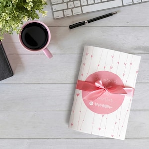 Puede incluir: Una tarjeta rectangular blanca con un patrón de líneas verticales y pequeños corazones rosas, envuelta en una cinta de satén rosa. Un círculo rosa central muestra el texto Te queremos y nosotros, junto a un gráfico de onda sonora musical blanco.