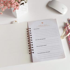 May include: A white spiral bound journal with a pink heart and the text "Reflection Diary" on the cover. The journal is open to a page with lined paper and the text "What sparked joy?" at the top.
