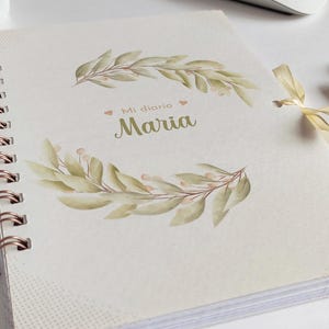May include: A white spiral bound journal with a gold coil. The cover features a watercolor illustration of green leaves and the text "Mi diario Maria".