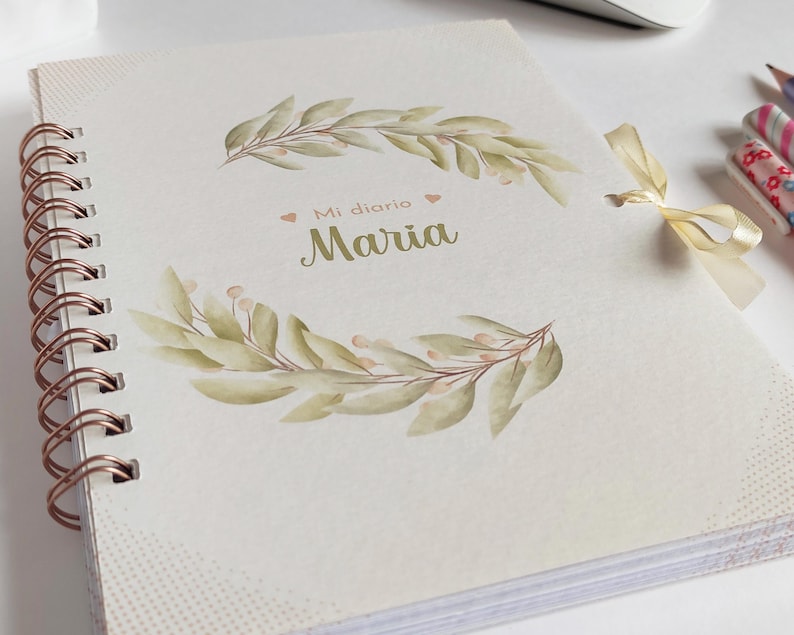 May include: A white spiral bound journal with a gold coil. The cover features a watercolor illustration of green leaves and the text "Mi diario Maria".
