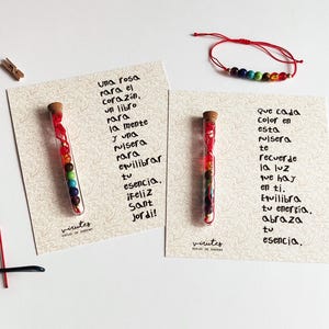 Puede incluir: Dos tarjetas con mensajes inspiradores y viales de vidrio coloridos llenos de cuentas. El texto de la tarjeta de la izquierda dice "una rosa para el corazón, un libro para la mente y una pulsera para equilibrar tu esencia. ¡Feliz Sant Jordi!" El texto de la tarjeta de la derecha dice "que cada color en esta pulsera te recuerde la luz que hay en ti. Equilibra tu energía. Abraza tu esencia."