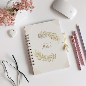 May include: A white spiral bound journal with a gold spiral and a floral design on the cover. The cover has the text "Mi diario Maria" in gold lettering.