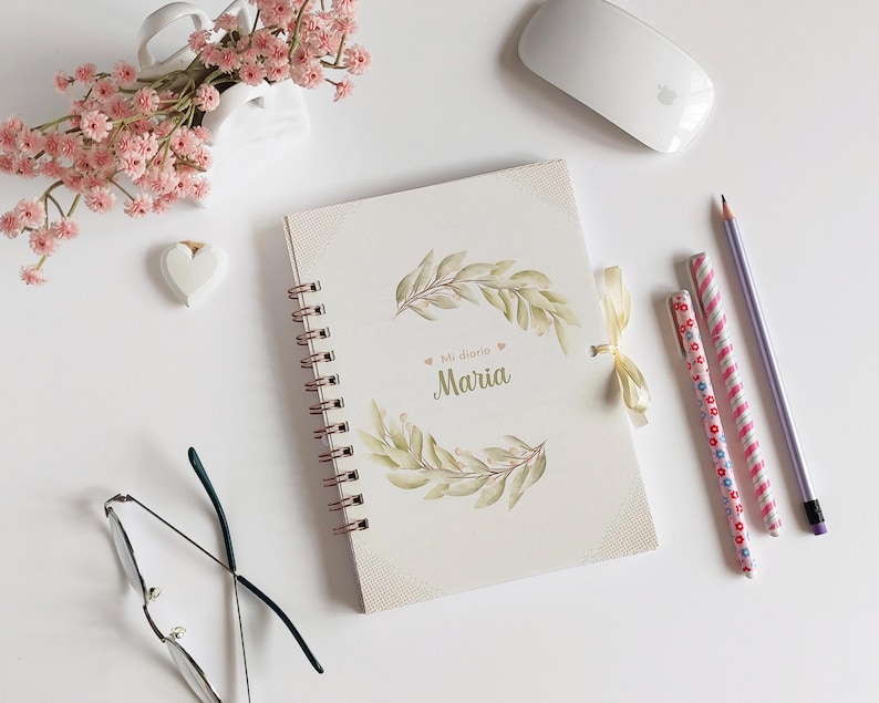 May include: A white spiral bound journal with a gold spiral and a floral design on the cover. The cover has the text "Mi diario Maria" in gold lettering.