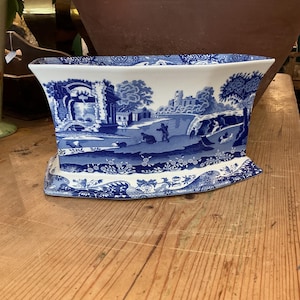 May include: A white ceramic planter with a blue transferware pattern featuring a landscape with ruins, trees, and figures. The vessel has a wide, flared rim and a rectangular base with rounded corners, decorated with intricate floral borders.