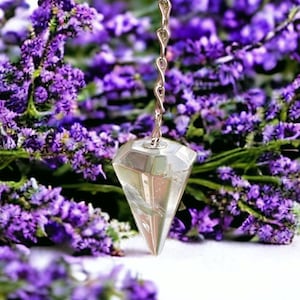 Op de afbeelding: Een helder kristallen slinger met een zilveren ketting die hangt boven een bed van paarse lavendelbloemen.