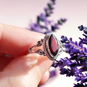 Könnte beinhalten: Ein silberner Ring mit einem roten Edelstein, der in einem filigranen, verzierten Design gefasst ist. Der Ring wird an einem Finger getragen und der Hintergrund ist unscharf mit lila Blumen.