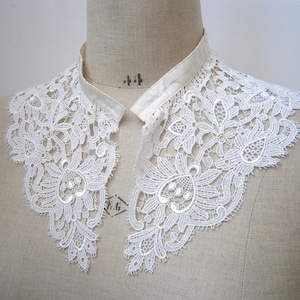 Puede incluir: Un cuello de encaje blanco con un intrincado dise&ntilde;o floral. El cuello est&aacute; hecho de encaje delicado y presenta un escote de cinta de sat&eacute;n. El cuello se exhibe en un maniqu&iacute;, mostrando su estilo elegante y vintage.