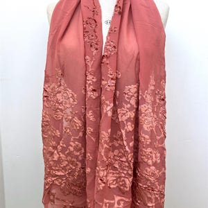 以下が含まれることがあります： 繊細な花柄刺繍が施された、コーラル色のシアーなスカーフ。マネキンにかけられ、繊細で流れるような生地と詳細なデザインを表現しています。花柄は縁と中央に集中しています。