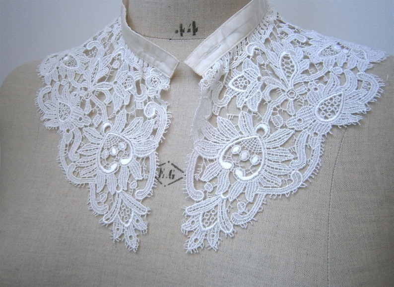 Puede incluir: Cuello de encaje blanco con dise&ntilde;o floral, expuesto sobre un maniqu&iacute;. El cuello presenta detalles intrincados y un delicado patr&oacute;n calado. Un accesorio de estilo vintage.