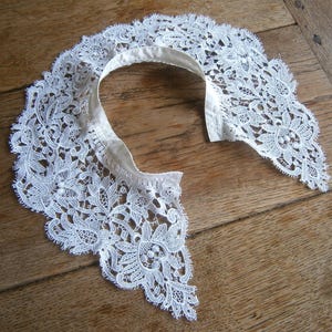 Puede incluir: Un cuello de encaje blanco con intrincados patrones florales y de hojas. El cuello tiene una banda de sat&eacute;n para el cuello y se muestra sobre una superficie de madera. El encaje es detallado y delicado, mostrando una est&eacute;tica vintage.